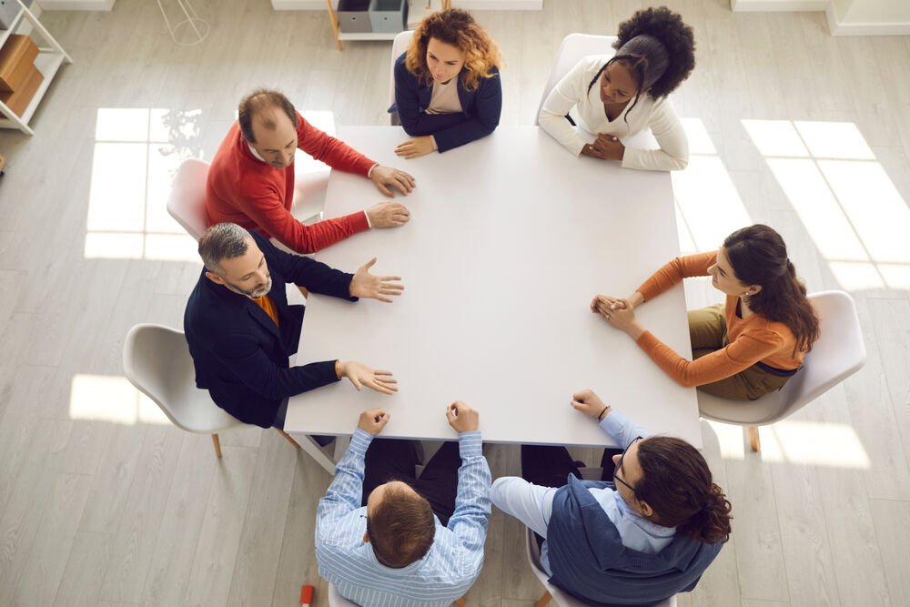 Group of diverse business people sitting around big modern square office table. High angle shot of team of mixed race company employees discussing work matters in corporate meeting